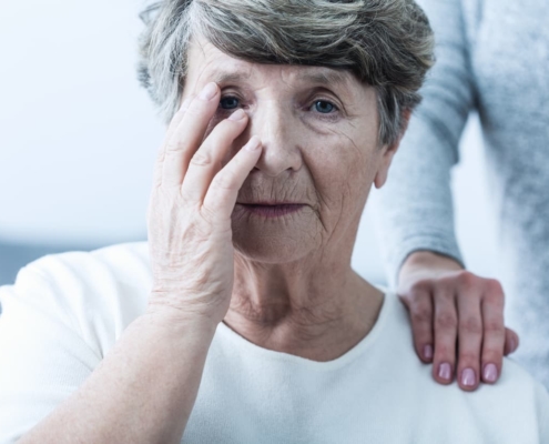 Elder abuse victim in nursing home covers face as a hand sits on her shoulder.