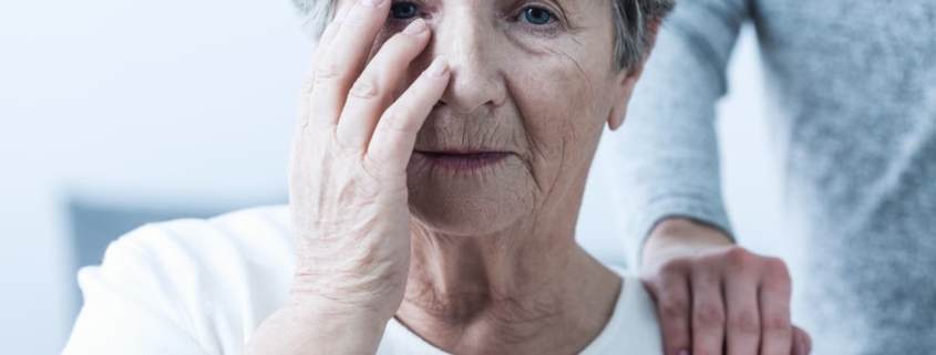 Elder abuse victim in nursing home covers face as a hand sits on her shoulder.