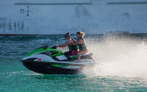 Two people riding a green and white jet ski, both wearing life vests, water spraying behind them, with a large ship in the background, text that reads: "TUG". common jet ski injuries