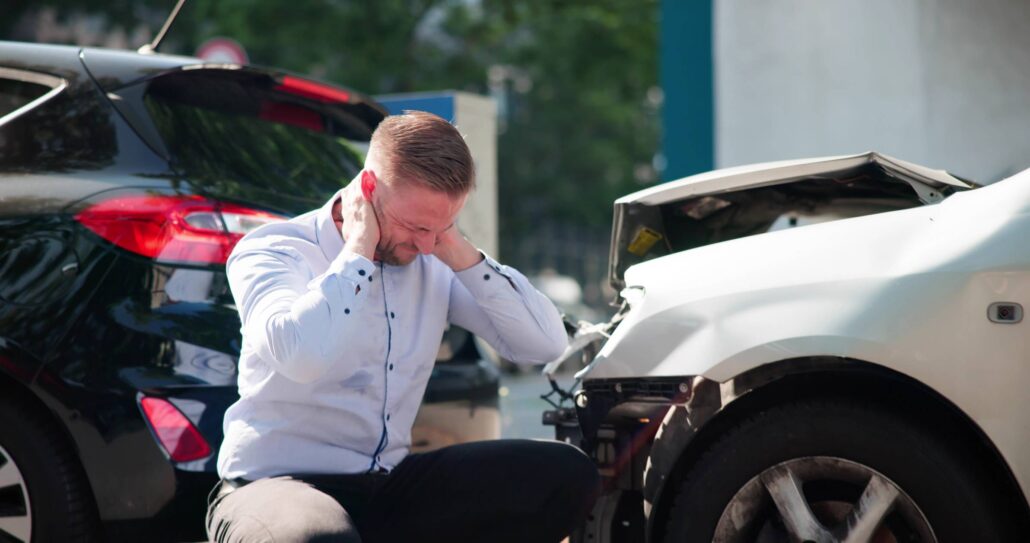 boca raton car accident lawyer - Man kneeling and holding neck in pain beside damaged cars after collision.