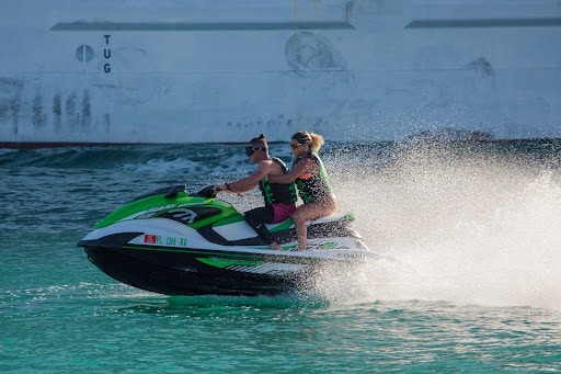 Two people riding a green and white jet ski, both wearing life vests, water spraying behind them, with a large ship in the background, text that reads: "TUG". common jet ski injuries